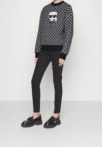 Schwarzer und weißer gemusterter Pullover mit einer Comicfigur in Sonnenbrille. Slim-Fit schwarze Hose und schwarze klobige Schuhe runden das Outfit ab.