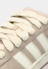 Sneaker in suede beige con strisce crema, lacci texture e un design cucito sottile. Suola in gomma con accenti lisci.