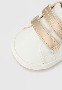 Λευκό συνθετικό παιδικό παπούτσι με μεταλλικές χρυσές λουράκια βέλκρο και κείμενο μάρκας, με στρογγυλεμένη μύτη και ομαλή υφή.