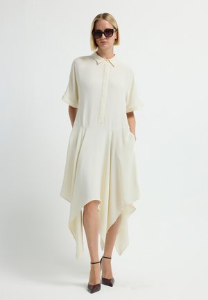 Licht crèmekleurige shirtdress met korte mouwen, knopen aan de voorkant en asymmetrische onderkant. Gemaakt met een paar donkere zonnebrillen en bruine puntige hakken.