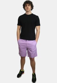 T-shirt nera a maniche corte, pantaloni corti lavanda con coulisse, sneakers nere con dettagli neon, in posizione eretta, sfondo neutro.
