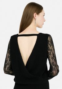 Top in maglia nera con profondo scollo a V sulla schiena e maniche lunghe in pizzo trasparente. Tessuto testurizzato con silhouette attillata e un piccolo dettaglio a taglio sulla parte posteriore.