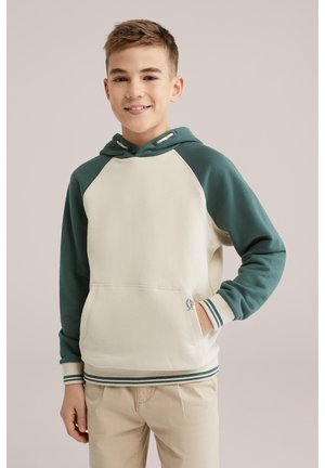 Pusaudzis zēns smaida, valkājot krēmkrāsas un zaļu kapuci ar kangu krūšu kabatu un bēšas krāsas bikses, stāvot pret vienkāršu fonu.