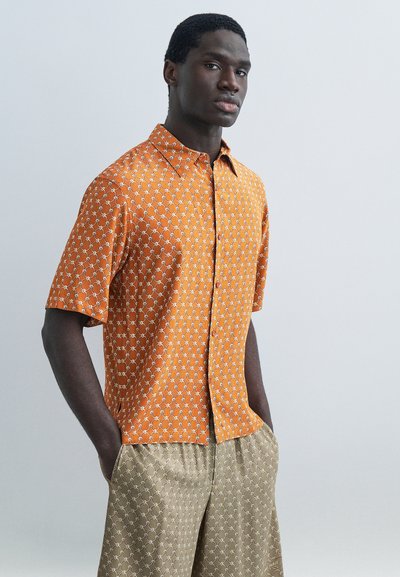 Homme portant une chemise à manches courtes orange à motifs et un short beige à motifs, les mains dans les poches, debout devant un fond uni.
