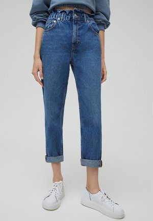 Personne portant un jean paperbag bleu taille haute roulé aux chevilles et des baskets blanches à plateforme, debout devant un fond clair uni.