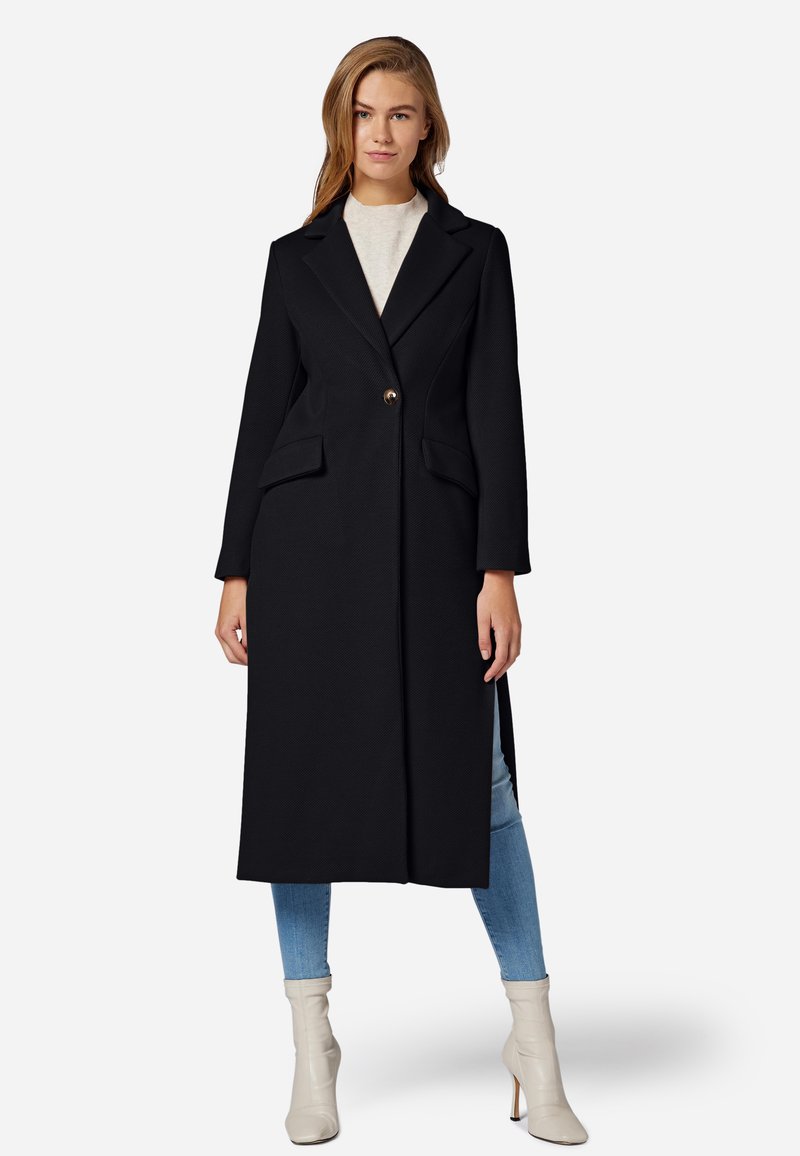 Schwarzer langer Mantel aus strukturiertem Stoff, mit einem Knopfverschluss und Seitentaschen. Das Model trägt hellblaue Jeans und cremefarbene Ankle Boots.
