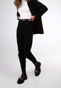 Pantalons noirs taille haute avec plis avant, associés à un haut ajusté à côtes blanches et des bottines noires à talon épais avec un détail en chaîne.