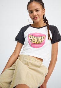 Tricou scurt cu mânecă scurtă, cu mâneci negre, corp alb, grafică cu o gură roz și textul "FLACKD"; asortat cu o fustă bej, până la genunchi.
