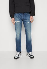 Vaqueros azules claros desgastados con desvanecimiento, roturas y diseño de pierna recta. Combinados con zapatillas deportivas negras y blancas.