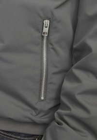 Gray bomber jacket featuring a zippered pocket with metallic hardware, smooth texture, and elastic cuffs. Close-up view of the design.