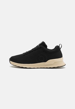 Zapatilla deportiva negra con parte superior de malla, punta redonda y suela beige. Presenta cordones, detalle de logo en relieve y un tirador en el talón.