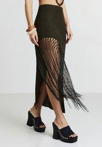 Gonna maxi verde oliva con dettagli in frangia laterale, spacco alto e tessuto testurizzato. Abbinata a sandali platform neri con borchie dorate.