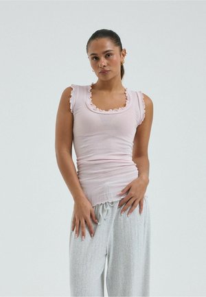 Femme portant un haut sans manches rose pâle avec des bords à volants et un pantalon gris clair à cordon, debout devant un fond uni.