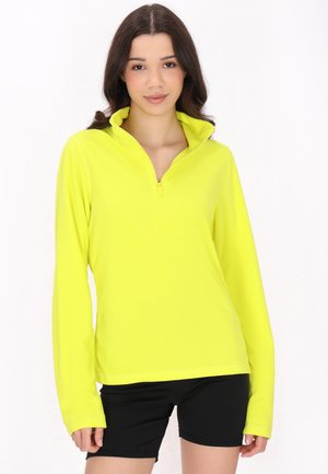 Lys gul langærmet pullover med en kvart lynlås, lavet af glat stof. Båret over sorte shorts med en afslappet pasform.