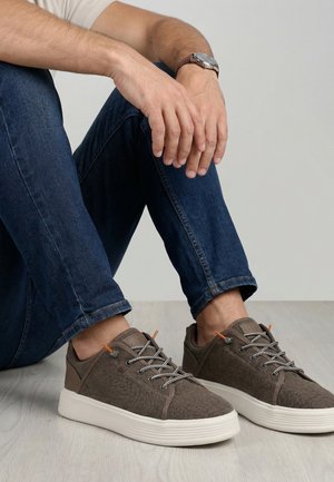 Muž sedící na podlaze, oblečený v tmavě modrých džínách a hnědých volnočasových teniskách nízkého střihu s bílými podrážkami a tkaničkami.