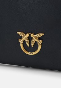 Sac à main noir texturé présentant un anneau doré orné de deux oiseaux en relief. Nom de la marque "PINKO" visible sur la quincaillerie.