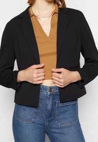 Blazer nero cropped con revers, realizzato in tessuto liscio, abbinato a un top marrone a coste e jeans blu a vita alta.