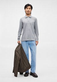 Polo grigio a maniche lunghe con colletto, abbinato a jeans di denim azzurro chiaro. Una giacca trapuntata verde scuro pende dal braccio sinistro.