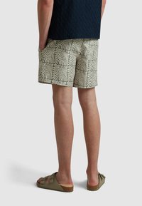 Groene korte broek met een wit bladmotief, voorzien van zijzakken. Gecombineerd met bruine sandalen op een neutrale achtergrond.