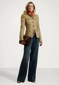 Šviesiai rudos lino švarkas su keturiais priekiniais kišenėmis, derinamas su tamsiai mėlynomis plačiomis kelnėmis. Modelis laiko rudą odinę rankinę ir vilki raudoną šaliką.