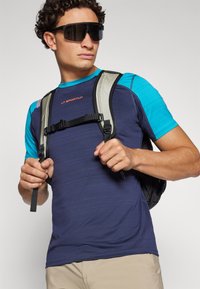 Ναυτικό μπλε αθλητικό μπλουζάκι με τιρκουάζ λεπτομέρειες, κοντά μανίκια και υφή, φορεμένο κάτω από μια γκρι και μαύρη τσάντα πλάτης. Σκοτεινά γυαλιά ηλίου.