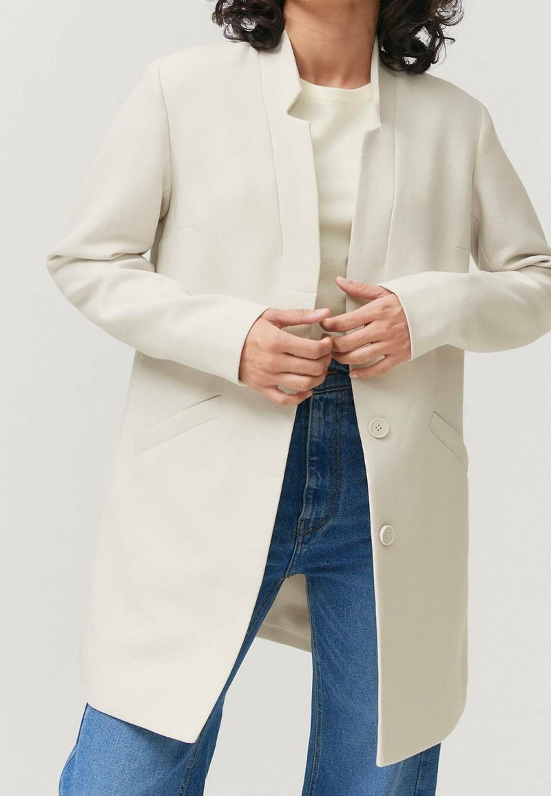 Abrigo beige de manga larga sin solapas, con dos bolsillos laterales y un cierre de botón único, llevado sobre una blusa clara y unos vaqueros.