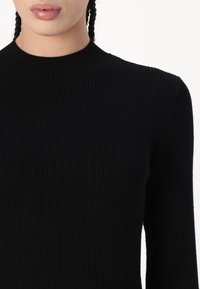 Top nero a coste con collo alto. Maniche lunghe e silhouette aderente, che mette in evidenza un tessuto testurizzato e una finitura liscia.