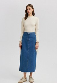 Blauwe denim maxi-jurk met subtiele versieringen, gecombineerd met een crèmekleurige top met lange mouwen en gefranjerde manchetten. Casual schoenen maken de outfit compleet.