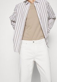 Camisa de botones a rayas en marrón claro y blanco, llevada sobre una camiseta beige, combinada con jeans blancos; ajuste casual y texturas suaves.