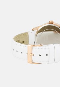 Valge nahkse kellarihma kinnitus roosa kullaga. Omab siledat tekstuuri ja peeneid õmblusi. Tagumine osa on roostevabast terasest, millel on graveeritud detailid.