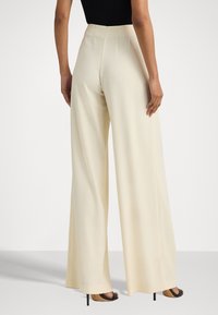 Nainen, joka on pukeutunut kermanvärisiin korkea vyötäröisiin väljiin housuihin ja mustiin korollisiin sandaaleihin, näkyy takaa valkoista taustaa vasten.