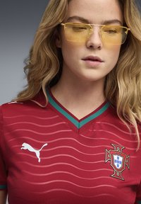 Κόκκινη φανέλα ποδοσφαίρου με υφασμένα ρίγες, πράσινες λεπτομέρειες στο V-λαιμόκοψη και λευκό λογότυπο Puma. Διαθέτει το έμβλημα της Πορτογαλίας στο στήθος.