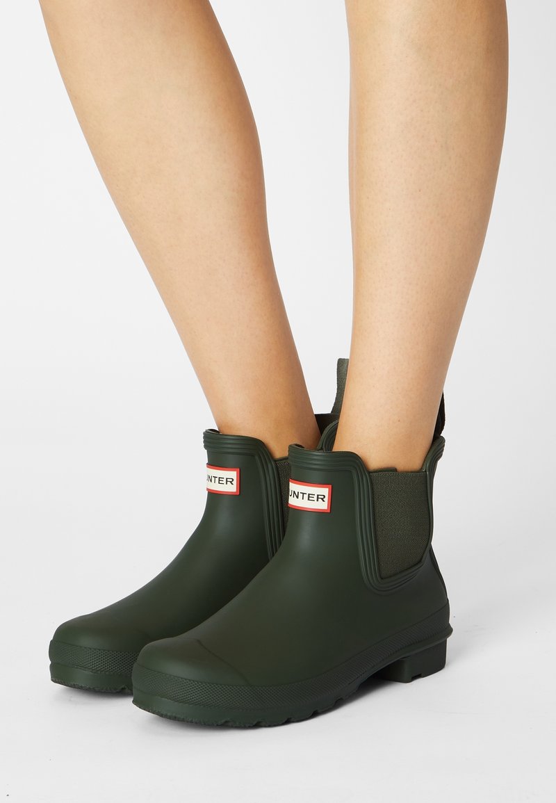 Chevilles et jambes inférieures portant des bottines en caoutchouc mat vert foncé avec des côtés élastiques et des logos rectangulaires blancs et rouges sur chaque devant.