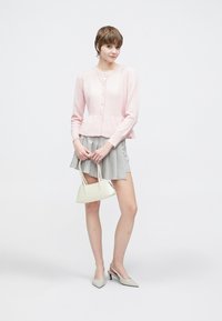 Jasnoróżowy dziergany kardigan z okrągłym dekoltem i zapięciem na guziki, zestawiony z szarą plisowaną spódnicą i białą torebką, dopełniony szarymi szpiczastymi szpilkami.