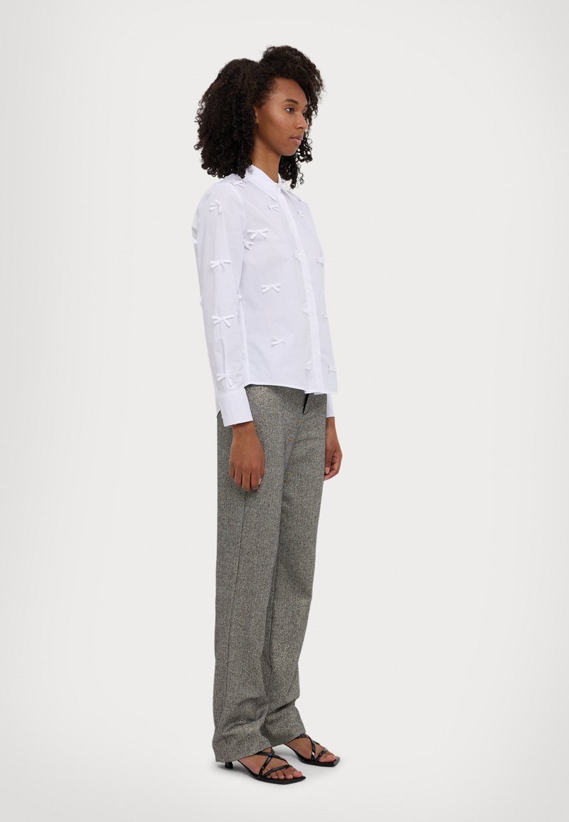 White shirt with bow details, paired with gray patterned trousers and black heeled sandals. The shirt features a button-down front and long sleeves.