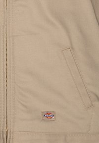 Beige work jacket made of sturdy fabric, featuring a gold zipper, a side pocket, and a Dickies logo patch on the lower section.