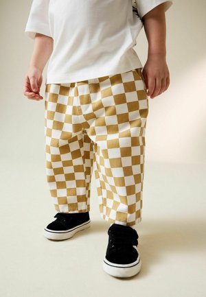 Gelbe und weiße karierte Baumwollhosen, kombiniert mit schwarzen Klettschuhen mit weißen Sohlen. Lässige Passform mit einem entspannten Design.