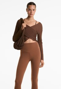 Smeđa rebrasta majica dugih rukava omotana oko struka, u kombinaciji s smešnim smešnim leggingama sa visokim strukom i mekom smeđom torbom za rame.