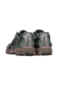 Scarpe da corsa verdi e grigie con parte superiore in rete, collarino imbottito e suola in gomma marrone spessa. Presentano il logo sul tallone e sul lato.