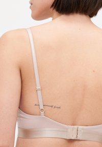Lys beige bh med justerbare stropper, glatt stoff og krok-og-løkke lukking. Har en synlig tatovering på ryggen.