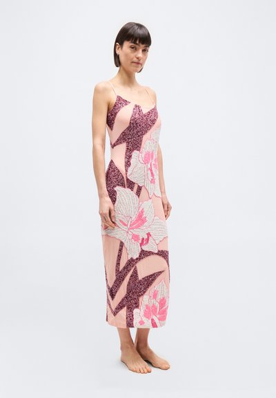 Γυναίκα που στέκεται ξυπόλητη φορώντας αμάν ικο ροζ φόρεμα με μεγάλα λουλουδάτα και γεωμετρικά γκλίτερ μοτίβα.