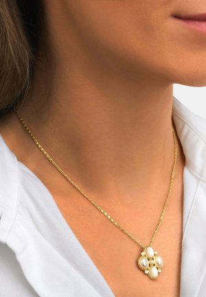 Collier chaîne en or avec un pendentif présentant quatre perles ovales disposées en forme de croix, porté au cou d'une femme sur une chemise blanche.