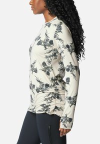 Frau mit langen lockigen Haaren, die ein beigefarbenes Langarmoberteil mit schwarzem Blumenmuster und schwarze Leggings mit einer Reißverschlusstasche trägt.