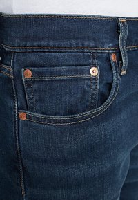 Jeans en denim bleu foncé avec rivets en cuivre, poches traditionnelles et coutures jaunes. Tissu texturé avec une ceinture standard.