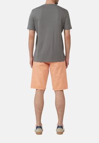 Graues Baumwoll-T-Shirt kombiniert mit hellorangefarbenen Jeansshorts. Das Model trägt beiges Schuhwerk mit blauen Akzenten. Sichtbare Nähte und Taschen an den Shorts.