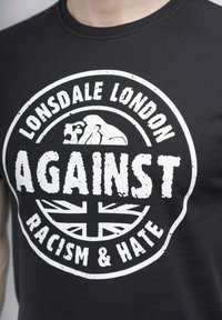 Juodas medvilninis marškinėliai su apvalia balta grafika, kurioje užrašyta "Lonsdale London" ir "PRIEŠ RASIZMĄ IR NEAPYKANTĄ", įskaitant Didžiosios Britanijos vėliavos dizainą.