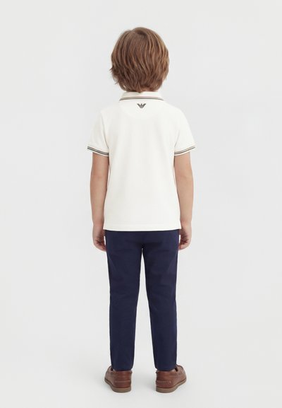 Παιδί που φοράει λευκό πόλο με κοντό μανίκι και λογότυπο στην πλάτη, μπλε ναυτικό παντελόνι και καφέ παπούτσια, στέκεται γυρισμένο με την πλάτη σε λευκό φόντο.