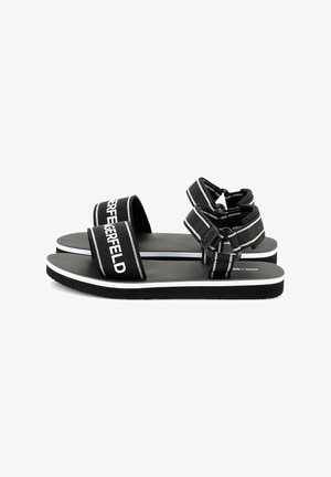 Svarta sandaler med två breda remmar som har vit text, platt sula och elastiska spännen. Texturerad fotbädd och kontrasterande vit kant.