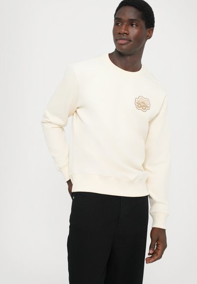 Lys kremfarget sweatshirt med rund hals, ribbestrikkede mansjetter og nederkant, med en teksturert logo-patch på venstre bryst. Brukt med svarte bukser.