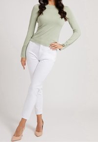 Pull léger vert à manches longues avec texture côtelée et détail de logo, associé à un jean blanc ajusté et des chaussures à talons nus.